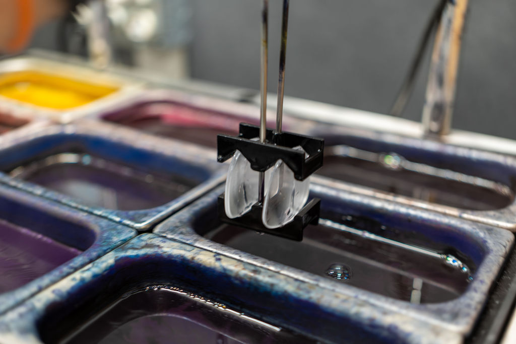 Close-up of clear lenses in a holder suspended above trays of yellow, purple, and blue lens dye